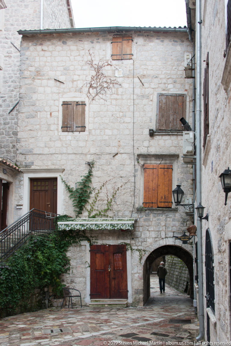 Pale stone courtyard façade with wooden shutters, doors, climbing vegetation, lamps, and an arched passage.