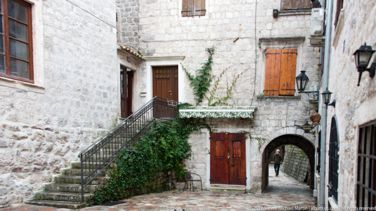 Stone courtyard with exterior stair, ivy, wooden doors, wall lamps, and a low arched passage in Kotor.