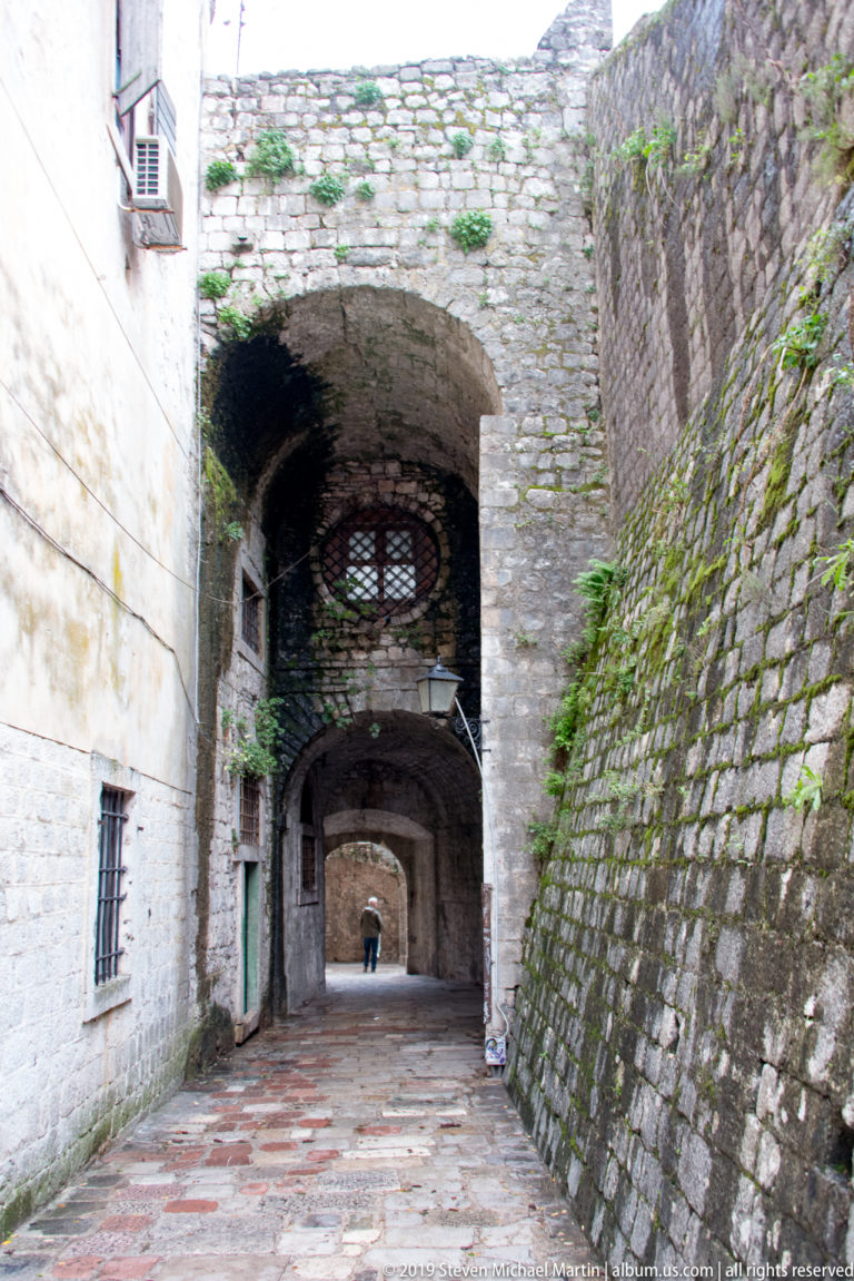 Long vaulted passage between thick stone walls with iron grilles, mossy masonry, and a distant person.
