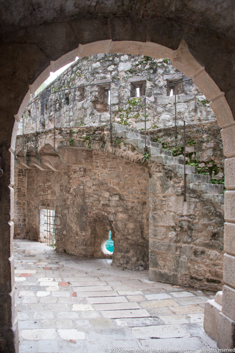 Broad stone arch framing a paved courtyard with weathered walls, railings, barred openings, and a small doorway.