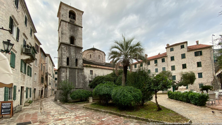 Stone church and tall square bell tower beside palms, shrubs, paving, and historic buildings in Kotor Old Town.