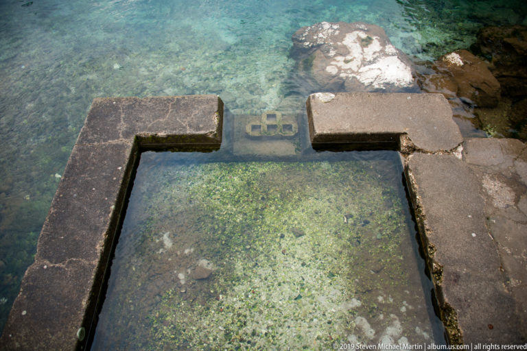 Rectangular stone-edged opening or basin beneath clear green water, with submerged gravel and masonry visible.