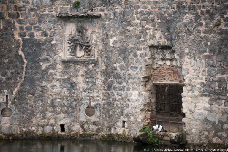 Weathered stone wall at the waterline with barred opening, small relief panel, and damp-stained masonry.