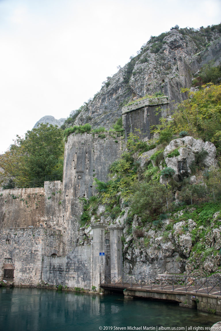 Stone defensive outwork at the waterline beneath a rocky, shrub-covered mountain slope.