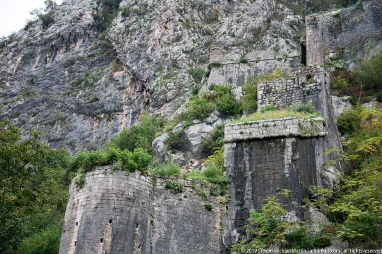 Ruined stone walls and tower-like defensive structures climbing a steep rocky slope above Kotor.