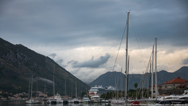 Sailboat masts and yachts in Kotor marina beneath dark clouds and steep mountains.