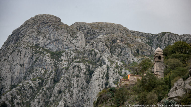 Small stone church tower below enormous gray limestone cliffs on Kotor’s mountain slope.