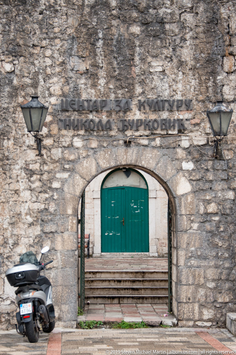 Arched stone gateway with Cyrillic lettering, two lanterns, green doors, and a parked scooter.