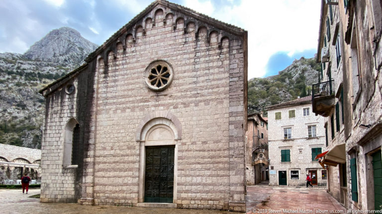 Small stone Church of St. Luke with rose window and round-arched doorway beneath steep mountains in Kotor.