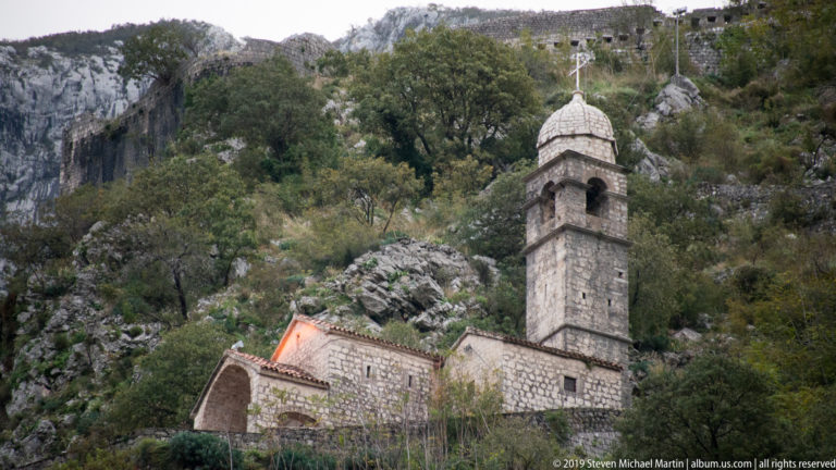 Stone church with narrow bell tower and orange roof set on a rocky mountain ledge above Kotor.
