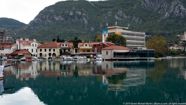 Waterfront buildings and moored boats reflected in calm water below steep green mountains.