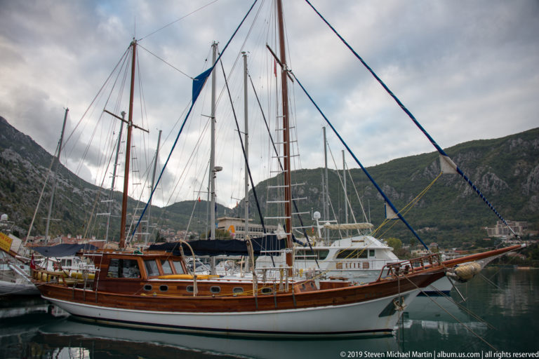 Large varnished wooden sailing vessel with tall masts and bowsprit moored in Kotor harbor.