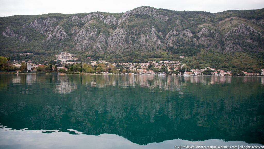 Wide bay view with shoreline houses and rugged limestone mountains reflected in turquoise water.