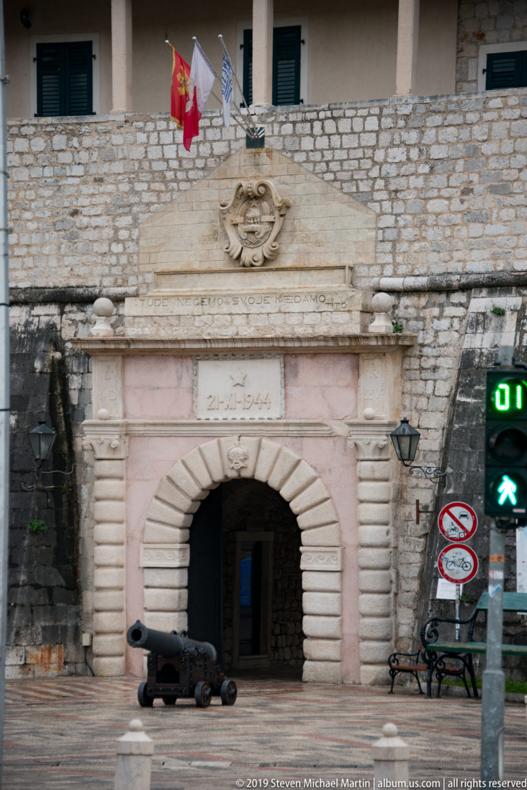 Monumental rusticated stone gate with carved relief, flags, cannon, and pedestrian signal at Kotor.