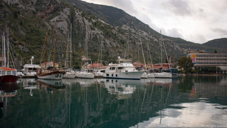 Sailboats and motor yachts moored in calm marina water beneath steep mountains at Kotor.