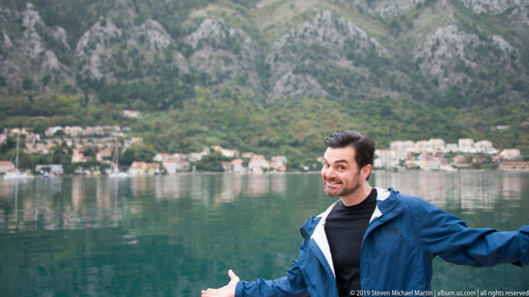 Man in blue jacket gesturing beside calm bay water with houses and steep mountains behind.