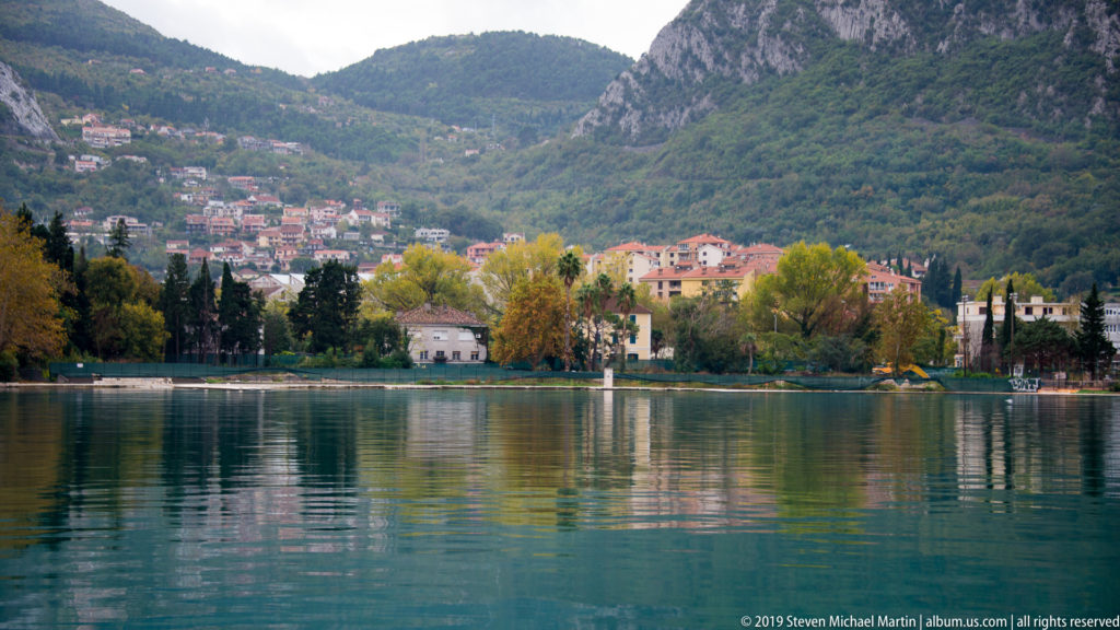 Waterfront houses and autumn trees reflected in calm bay water below steep forested mountains.