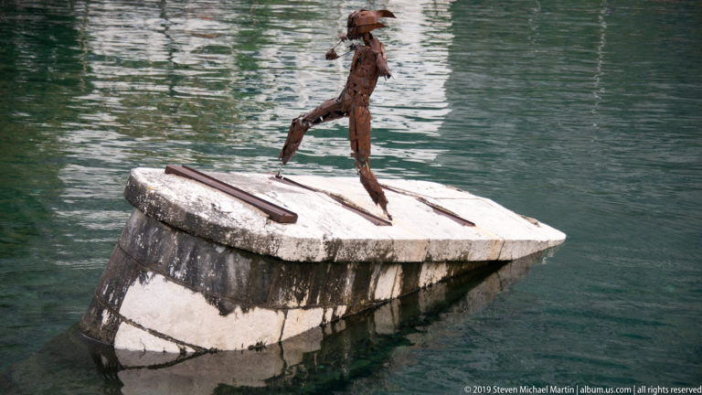 Rust-colored metal figure standing on a pale, partially submerged stone block in green water.