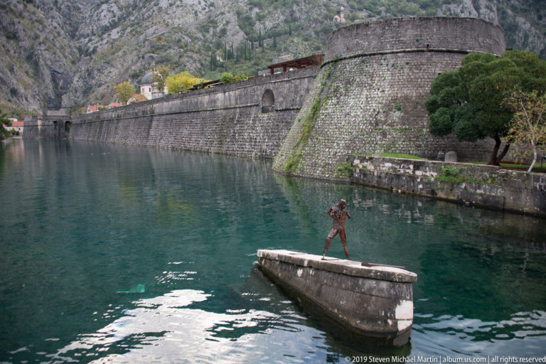 Long stone fortification wall beside calm green water, with a person standing on a small platform in front.