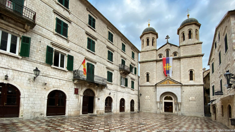 St. Nicholas Church with twin domed towers facing a patterned stone square beside a long shuttered building.