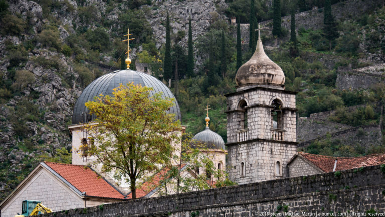 Large dark church dome, smaller dome, and stone bell tower with Orthodox crosses against a rocky slope.