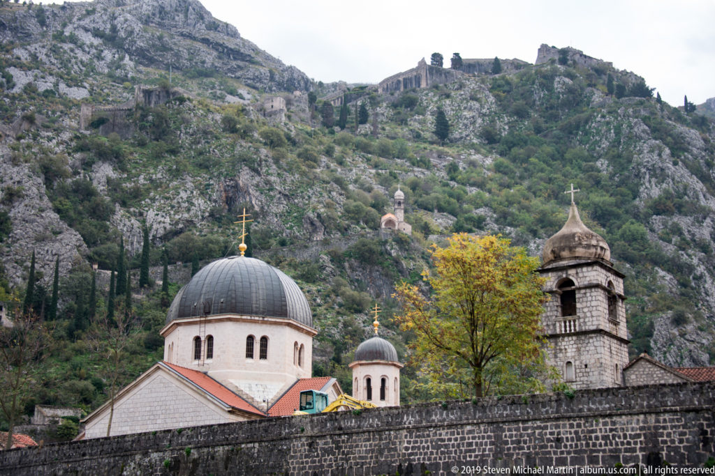 Orthodox church domes and bell tower below a rocky mountain slope with fortification remains above.