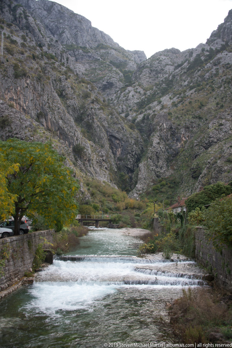 Fast-flowing stream crossing low weirs below steep gray limestone mountains near Kotor.