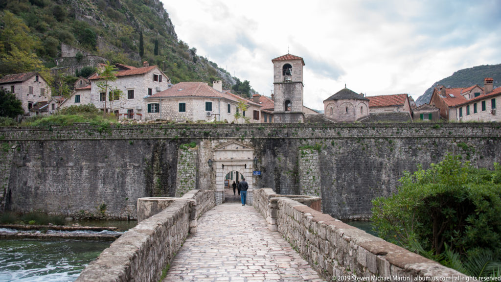 Centered bridge leading to a gate in Kotor’s wall, with houses and a church tower against the mountain.