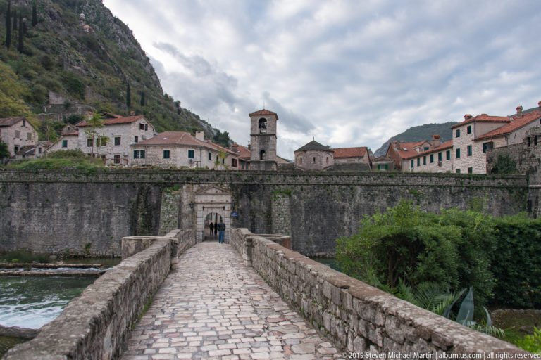 Cobblestone bridge leading to an arched gate in the wall, with church tower and red roofs beyond.