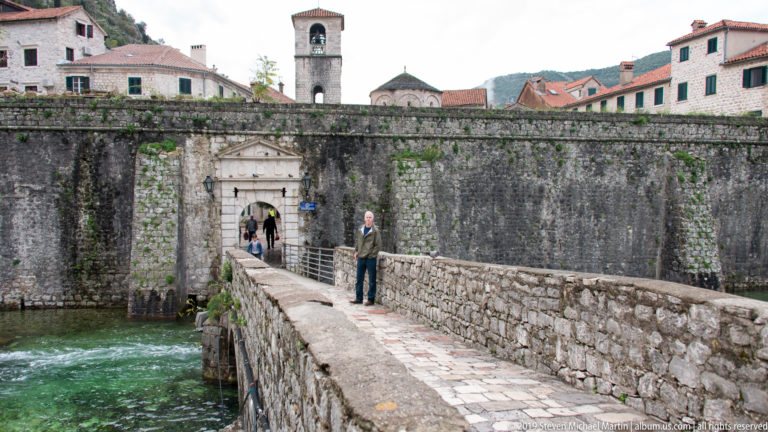 Stone bridge leading over green water to a gate in the city wall, with a bell tower rising behind.