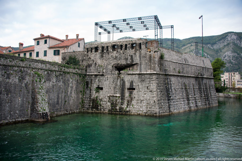 Massive stone corner bastion at the waterline with old-town buildings and mountains behind.