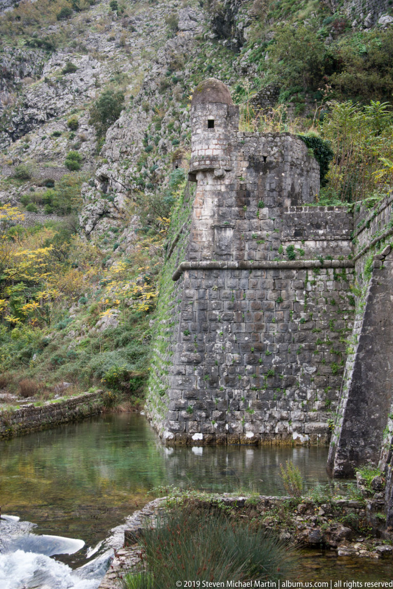 Angular stone bastion rising from green water below a steep rocky mountain slope.