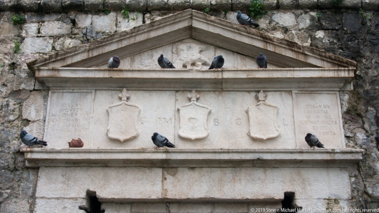 Close detail of a triangular stone pediment with carved shields, inscriptions, ledges, and several perched pigeons.
