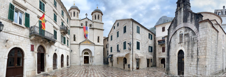St. Nicholas Church with twin domed towers and flags at the end of a narrow paved space in Kotor.