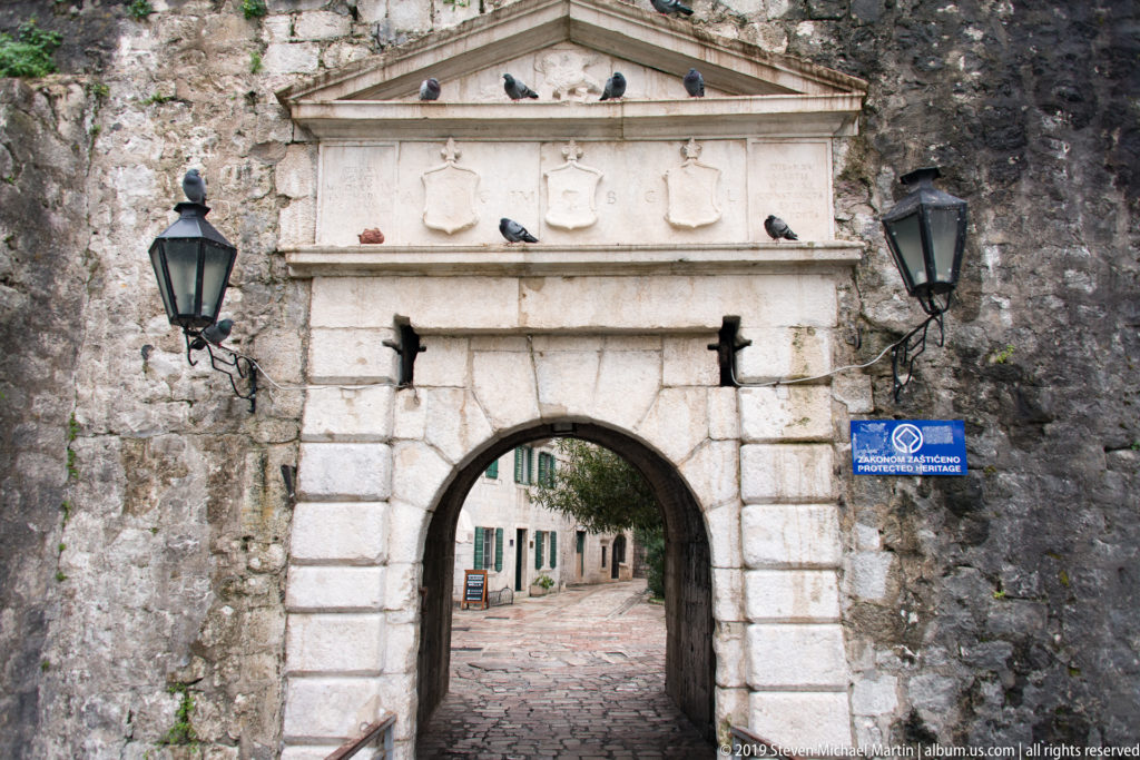 Arched stone city gate with pediment, carved shields, lanterns, and a paved lane visible through the opening.