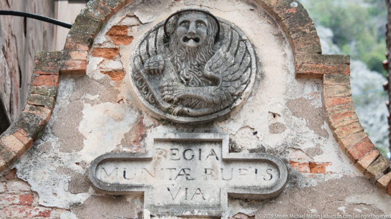 Weathered round relief of a winged lion above a carved Latin inscription in patched stone and brick masonry.