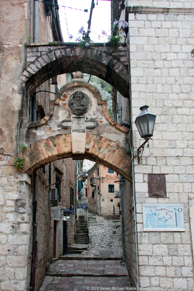 Stone archway with a carved crest above frames a narrow stepped lane in Kotor Old Town, beside a map sign and lamp.