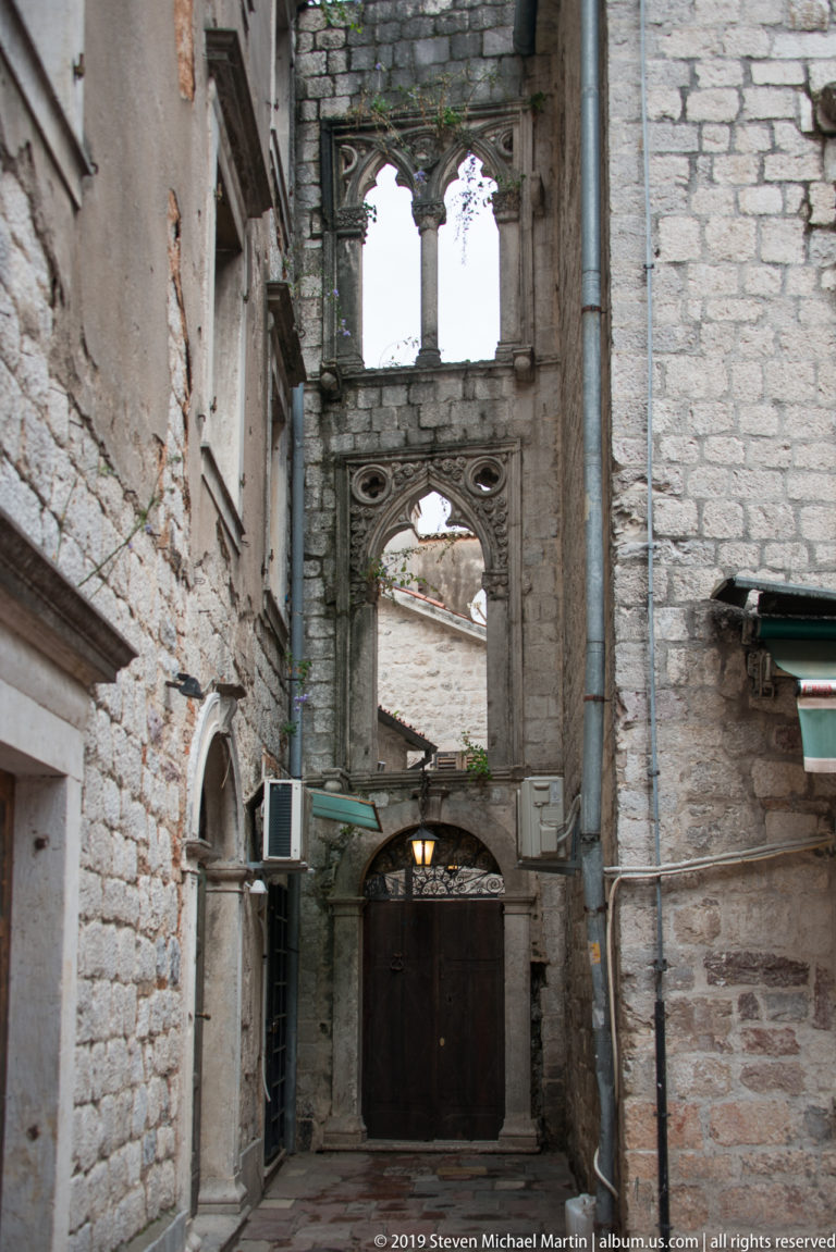 Narrow stone gateway with ornate carved openings and an upper loggia, surrounded by pipes and old masonry in Kotor.