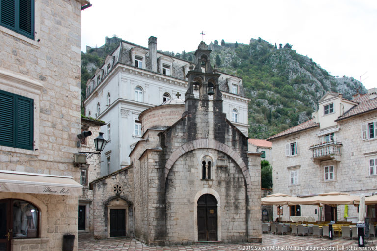 Small stone church with a bell gable and arched doorway facing a café-lined square in Kotor Old Town.