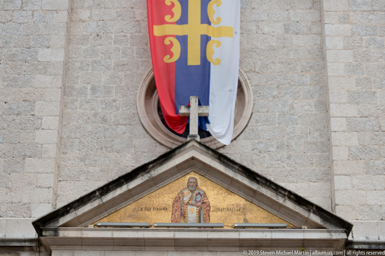 Triangular mosaic of a haloed bishop-saint above a cathedral doorway, with a large red, blue, and white banner overhead.