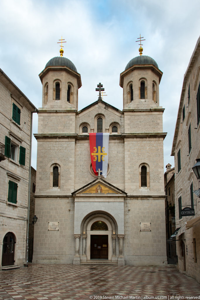 Front façade of Saint Tryphon Cathedral with twin towers, central doorway, pale stone walls, and a banner above the entrance.