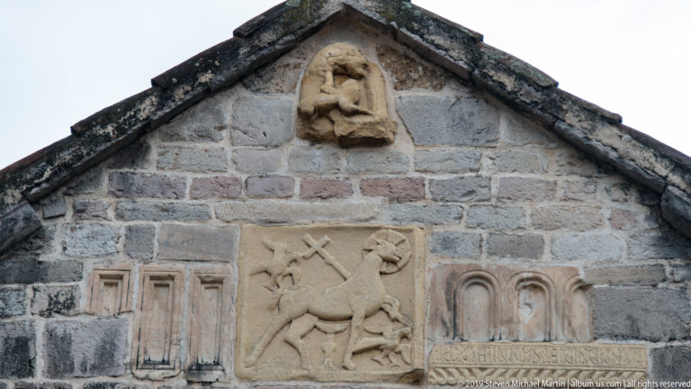 Weathered stone reliefs on a church gable, including a central Lamb of God panel and a small seated figure above.