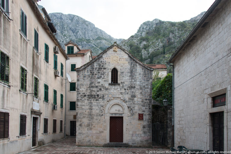 Front view of a small gabled stone church with arched door and window, tightly framed by taller Old Town buildings.