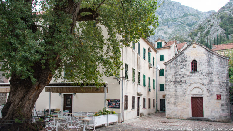 Small gabled stone church with an arched doorway and narrow upper window beside trees, café seating, and old town buildings.