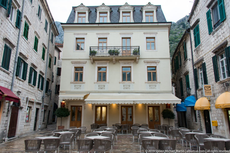 Pale historic building with balcony and dormers facing rows of café chairs in a small Kotor square.