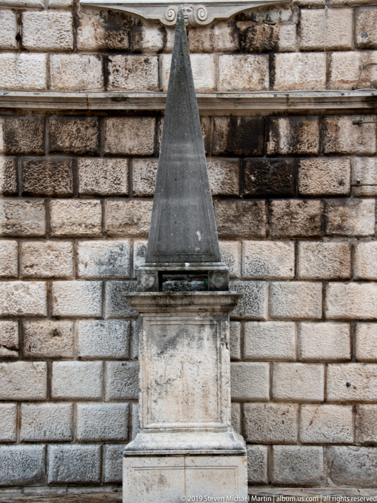Small dark stone obelisk on a stepped rectangular base positioned directly against a large block masonry wall.