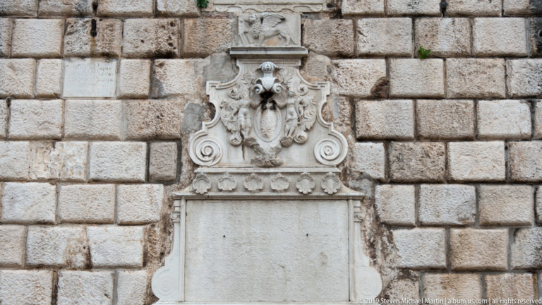 Tall carved stone crest with an oval shield and decorative scrollwork above a pale rectangular tablet on a rusticated wall.