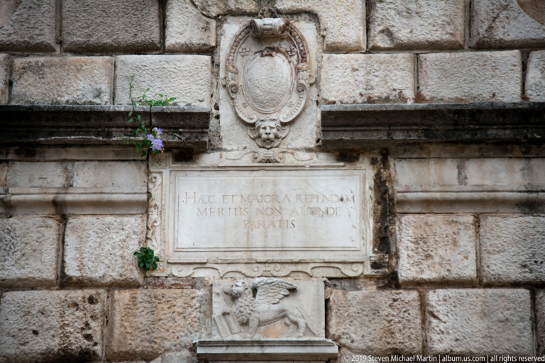 Inscribed stone plaque framed by an oval crest above and a winged lion relief below on a rusticated wall.