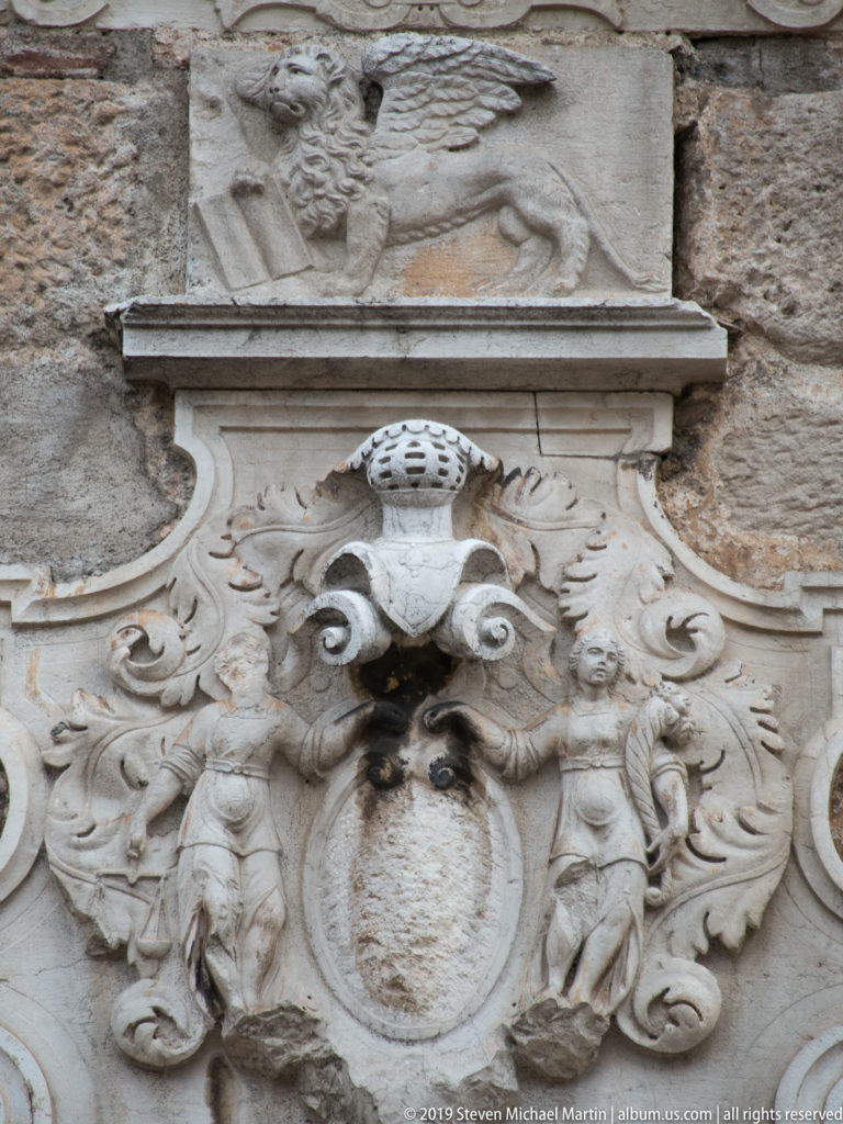 Ornate stone cartouche with scrolls and a central oval set below a relief of a winged lion on a masonry wall.