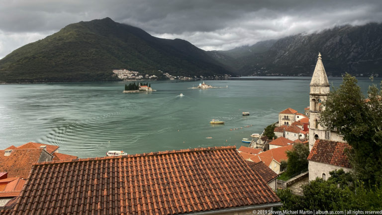 Red-tiled roofs and a pointed bell tower overlook the Bay of Kotor, with island churches and dark mountains beyond.